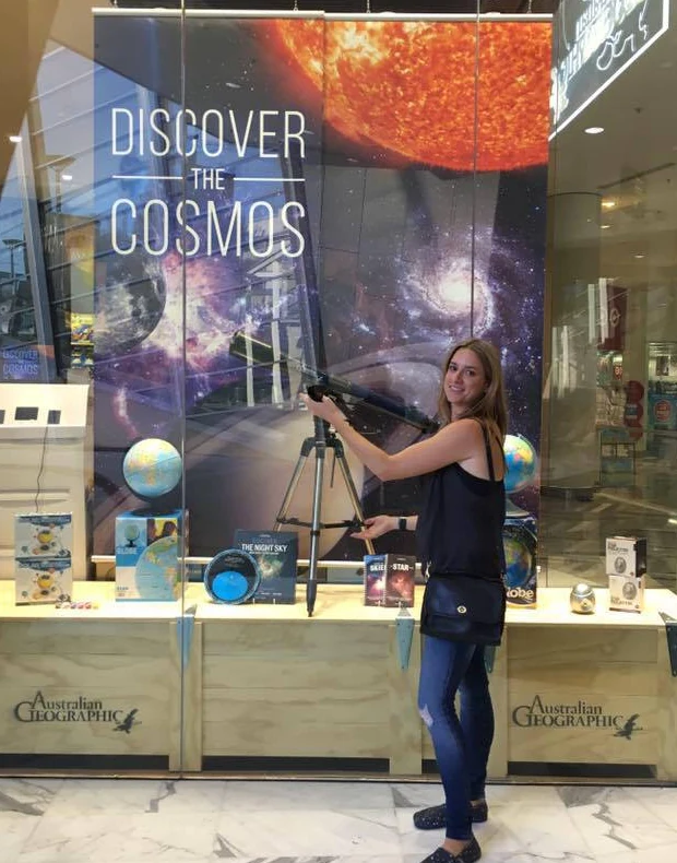 Anna Rosen beside a shop window display headed “Discover the Cosmos”, adjusting a telescope on a tripod in front of a backdrop of the Sun and a spiral galaxy.